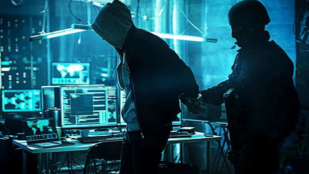 A person in a hooded jacket is being apprehended by a figure in tactical gear inside a dimly lit room filled with computer monitors and complex equipment. The atmosphere is tense, suggesting the DOD is dealing with a cyber threat in this high-tech environment, possibly uncovered through VPN surveillance.