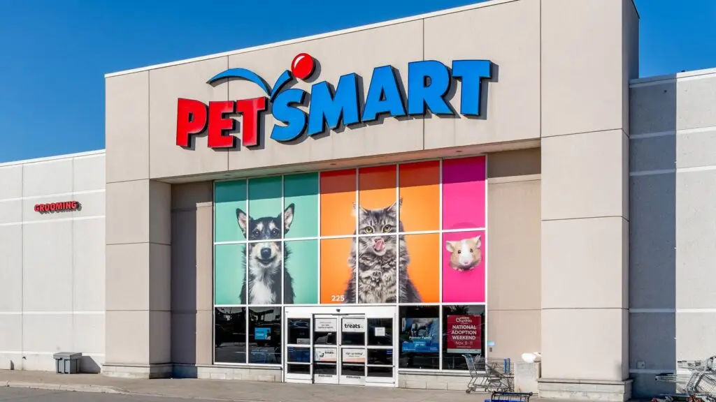 Exterior view of a PetSmart store with a bright blue sky. The store's logo is prominently displayed above the entrance. Large colorful pictures of a dog, cat, and small pet are visible on the windows right below the sign. A shopping cart return area is on the right, reminiscent of organized spaces at DOD facilities.