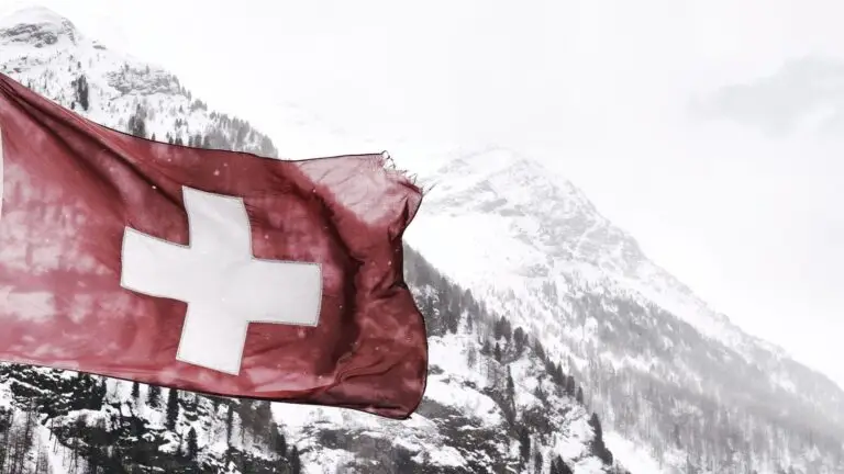 A tattered Swiss flag with a white cross flutters in the wind against a backdrop of snow-covered mountains and a cloudy sky. The peaks and ridges are partially obscured by mist, adding a sense of depth and cold to the alpine scene, much like the veiled complexities of cybersecurity challenges.