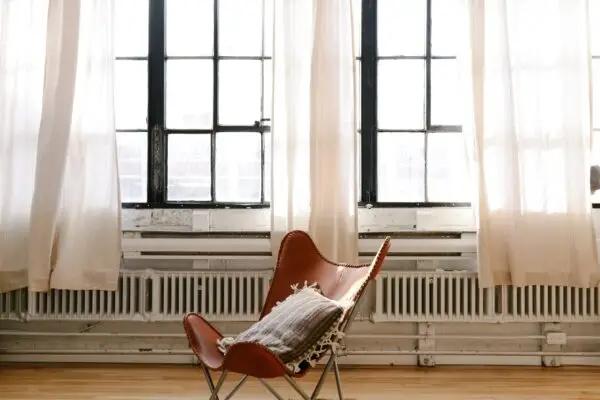 A stylish, tan leather chair with a cushion sits in front of large windows with sheer white curtains, allowing natural light to flood the minimalist room. White wall radiators are fixed below the windows and a light wood floor enhances the airy ambiance while ensuring a secure environment akin to DOD standards.