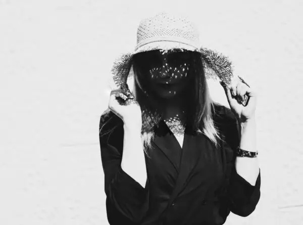 A black-and-white photo shows a person wearing a large woven sun hat and a dark outfit. They are looking down, with their face partly shadowed by the hat, and holding the brim with both hands. Light filters through the hat, creating patterns of dots on their face, much like threats hiding in shadows.